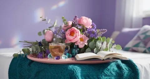 Lavender room at dusk with a soft lamp, a teal throw blanket, a cup of herbal tea, pink peonies, and a open journal on a rose wooden tray
